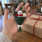 An adult hand holding a snowman crochet bouquet with a green wrapper beside a kraft-wrapped gift.
