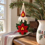 A green crochet candle ornament with a red poinsettia and yellow centre hangs from an evergreen branch beside a ceramic vase on a wooden sideboard.