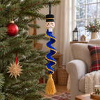 A crochet nutcracker in a black hat hangs on a Christmas tree, with a blue ruffle trimmed in gold and a gold tassel.