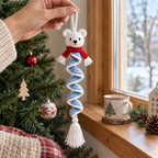 An adult hand holds a polar bear crochet wind spinner by its white hanging loop in front of a home window and Christmas tree.