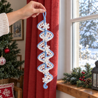 An adult hand holding the pale blue yarn loop of a white snowflake crochet lace spiral beside a red curtain and daylight window.