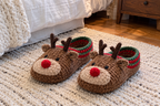 Reindeer crochet slipper pair: brown uppers with tan muzzles, red pompom noses, two short antlers, ears and red-and-green cuffs, on a bedroom rug.