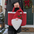 A woman in a winter coat carries a red gnome crochet tote beside a wreath-decorated door. A white looped beard hangs below the bag and a red cone hat folds over its handle.